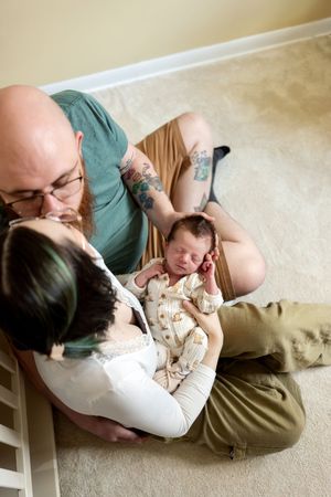 New parents sitting on floor together lovingly gazing at their sleeping newborn baby from above.