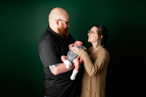 Happy new parents facing each other holding their newborn baby against a deep green background.