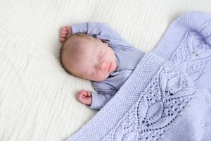 Sleeping newborn baby in blue outfit covered with a light blue knitted blanket.