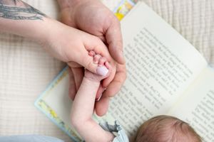 Adult hands gently holding a newborn baby's tiny hand near an open book.