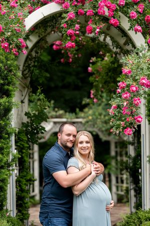 Couple embracing under a floral arch with pink roses during a maternity photo session in a lush garden.