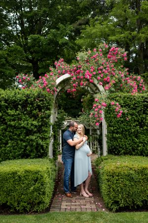 Expectant couple sharing a tender moment under a rose-covered pergola arch in a manicured garden.