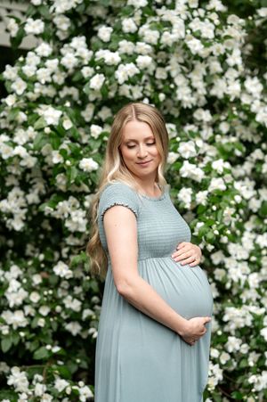 Pregnant woman in a sage green dress cradling her baby bump in front of white flowering bushes.