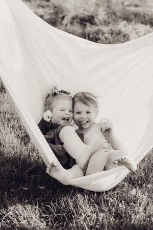 Two laughing young children cuddling together in a white fabric hammock outdoors.
