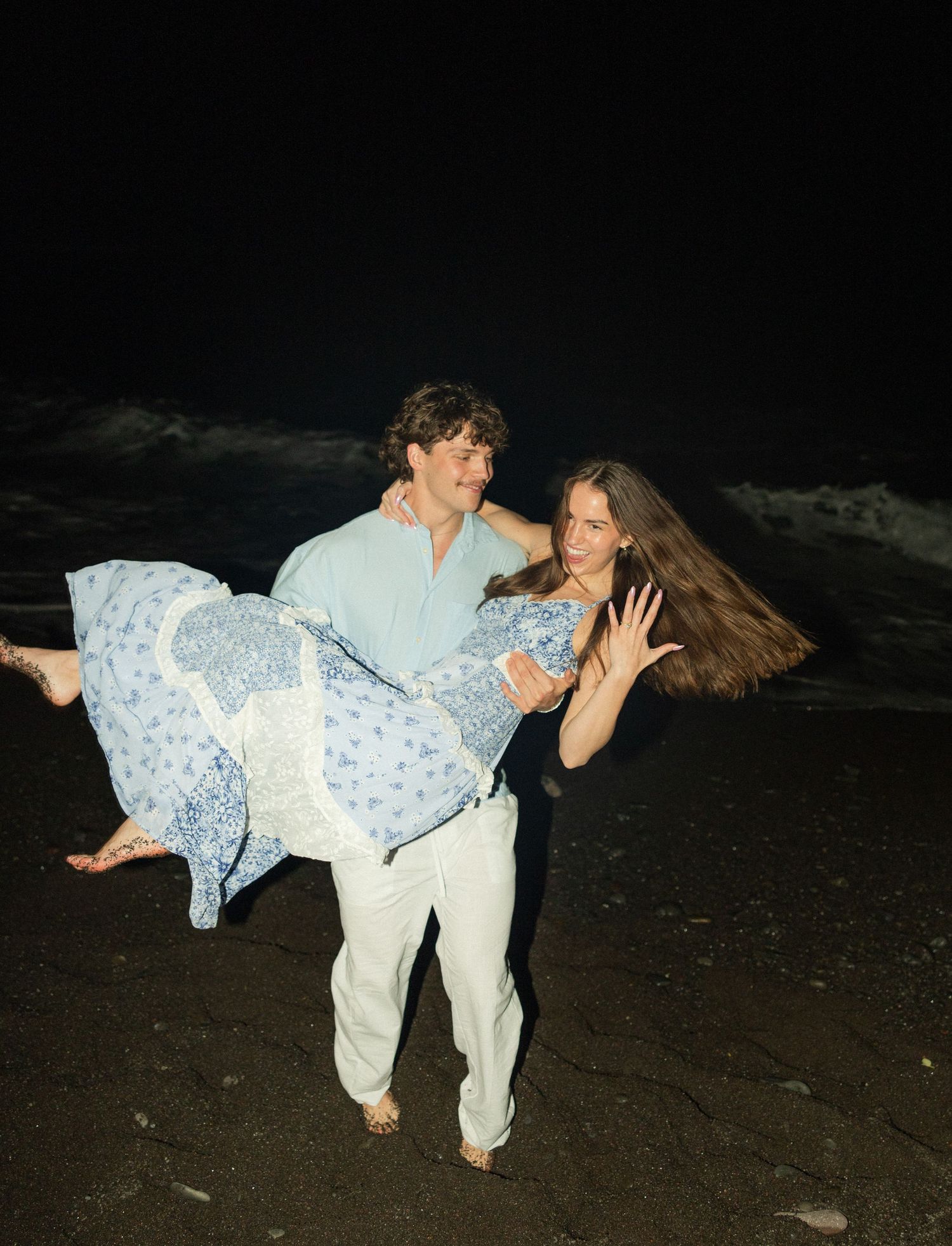 A couple shares a playful moment at night on the beach as one carries the other wearing a flowing blue floral dress.