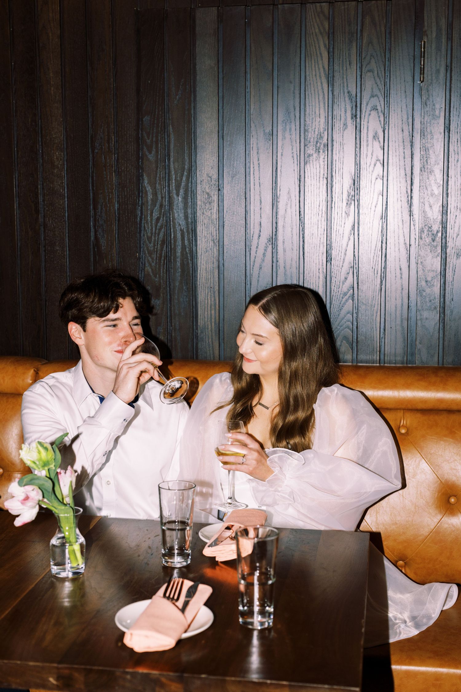 A couple in light-colored outfits shares drinks at a wooden table with a vase of flowers in a cozy booth setting.