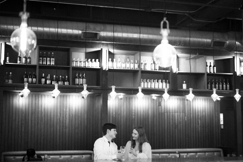 Two people enjoying drinks at a bar with glass pendant lighting and bottle displays on shelves.