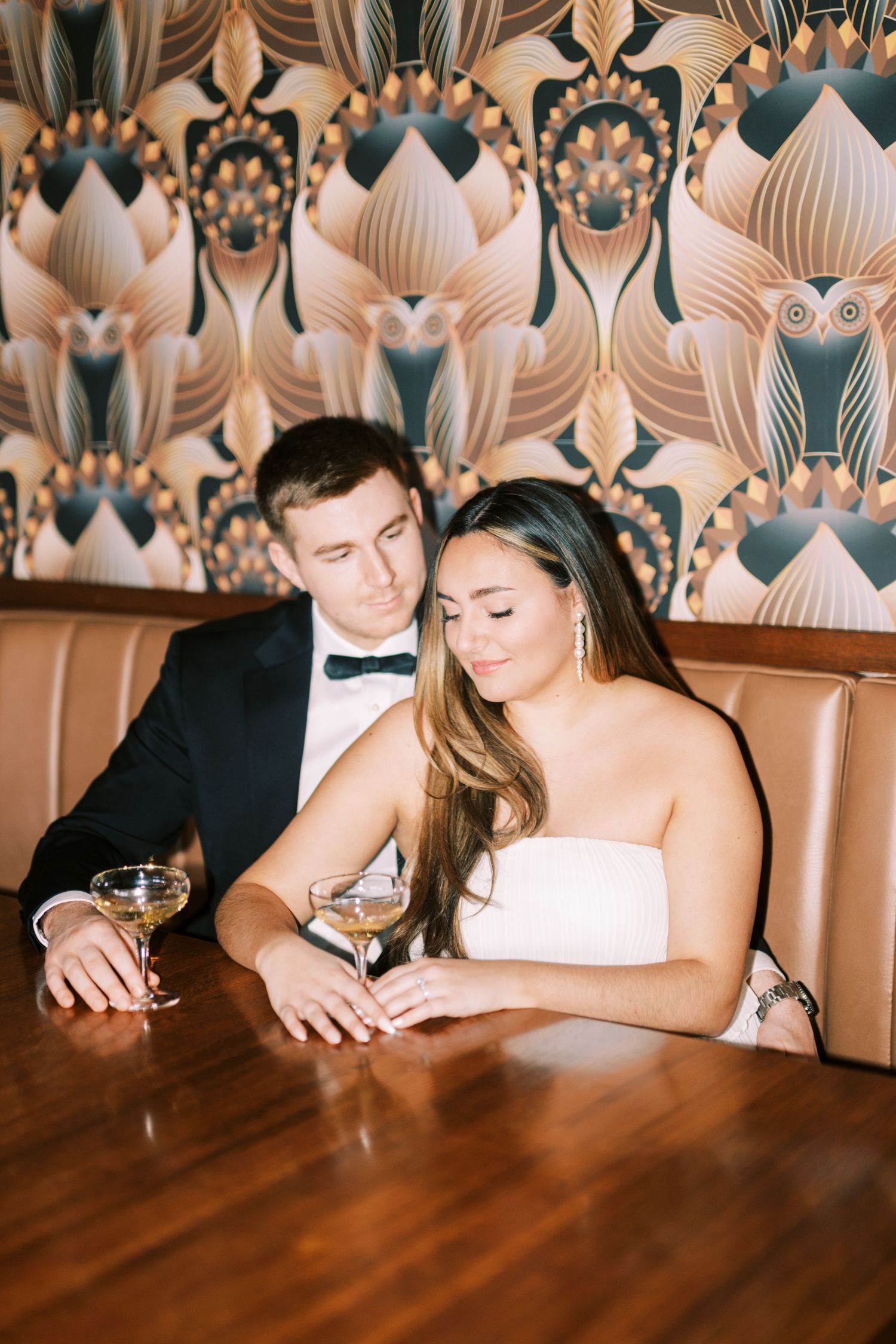 A couple sits intimately at a table with drinks, against a floral-patterned wall in a dimly lit restaurant setting.