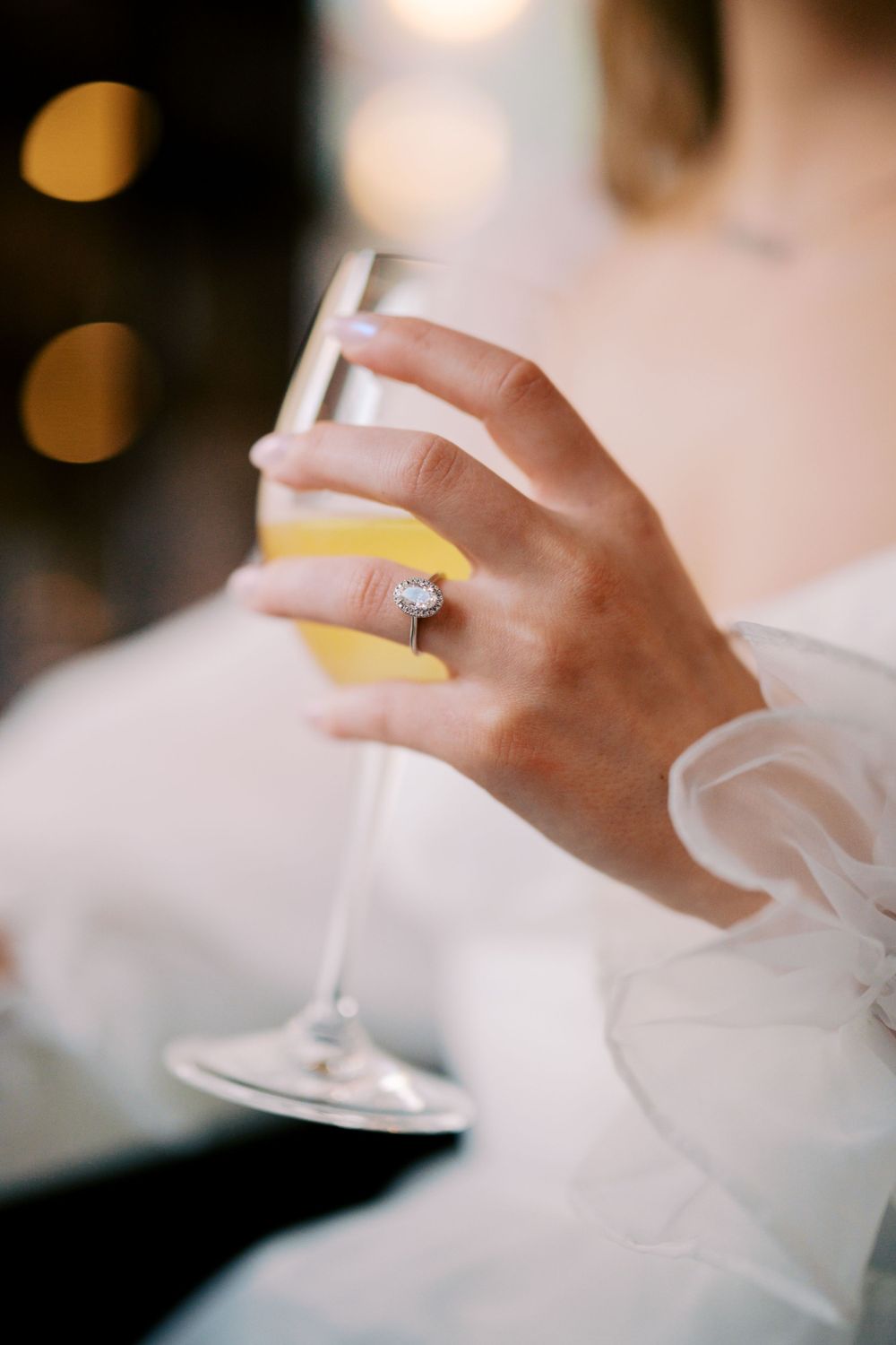 A hand adorned with a sparkling ring holds a champagne flute filled with a pale yellow drink.