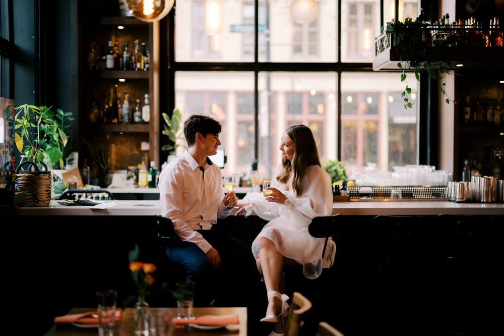 A couple enjoys drinks and conversation at a stylish bar with large windows.