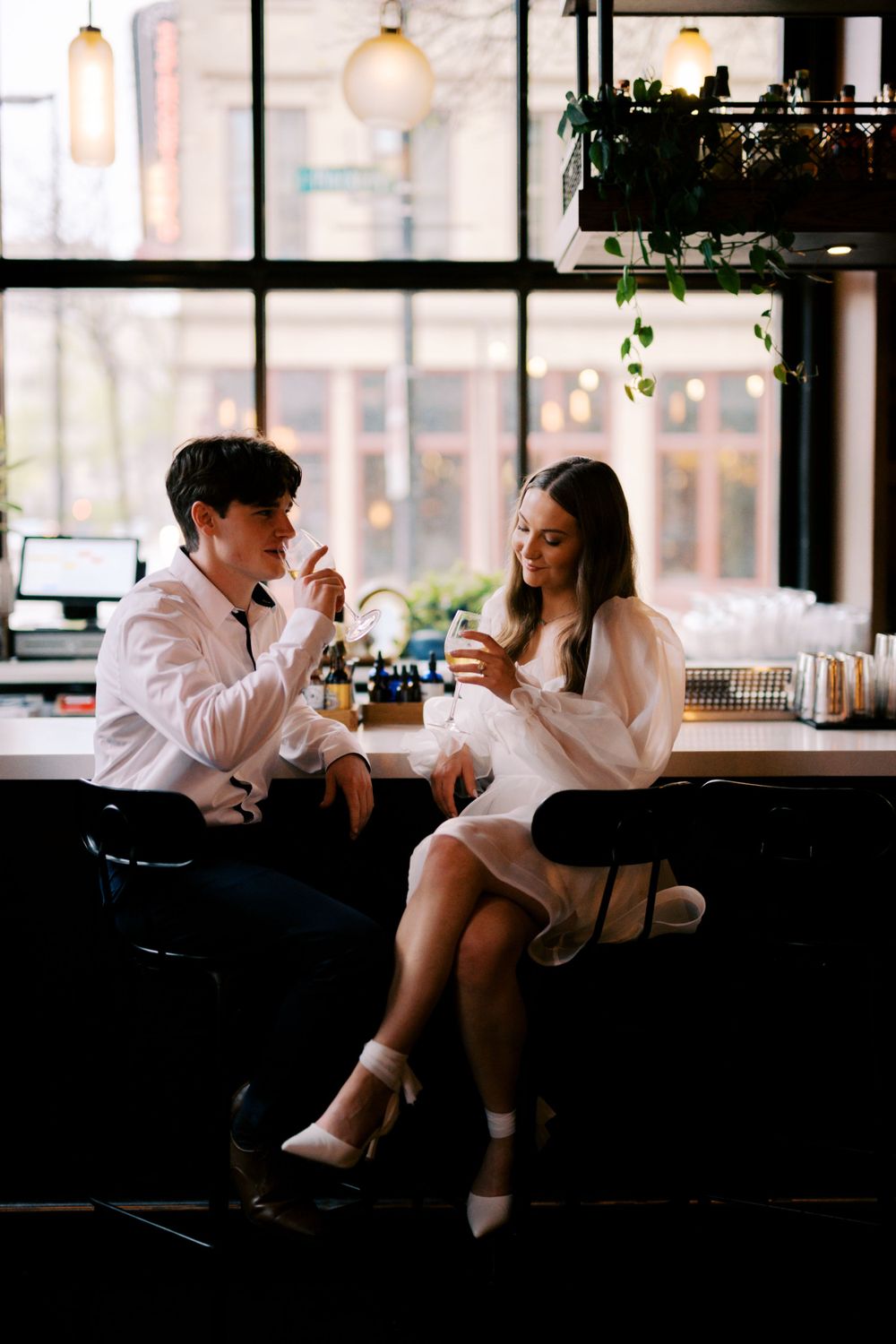 Two people sit at a bar counter, enjoying drinks in a cozy cafe with large windows and hanging plants.