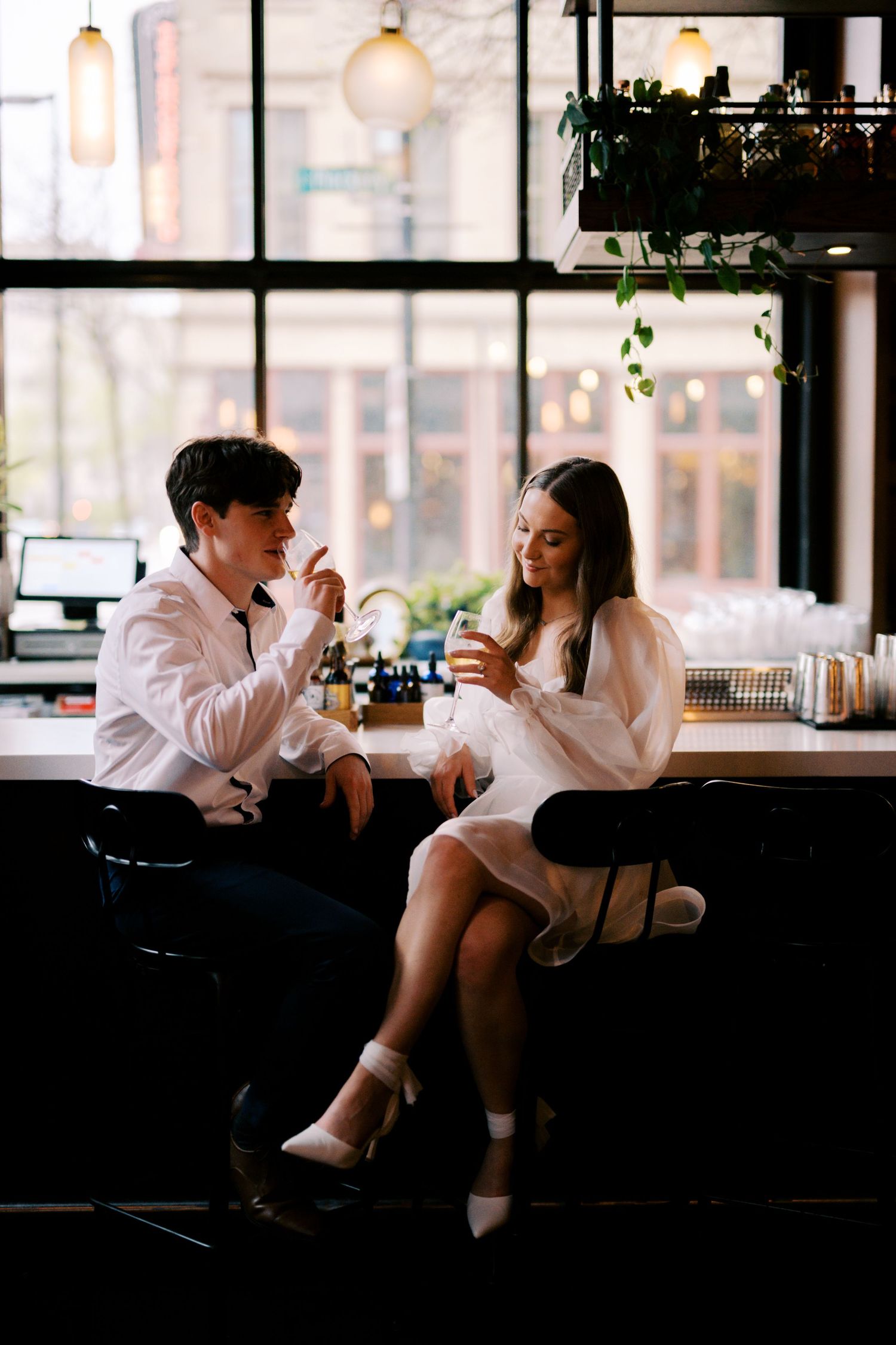 Two people sit at a bar counter, enjoying drinks in a cozy cafe with large windows and hanging plants.