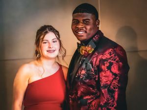 A couple in formal wear smiles together, with red one-shoulder dress and floral jacket.