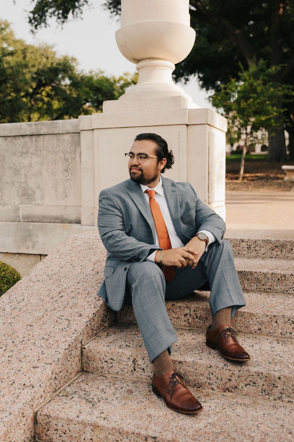 A person in a gray suit and orange tie sits relaxed on stone steps outdoors.