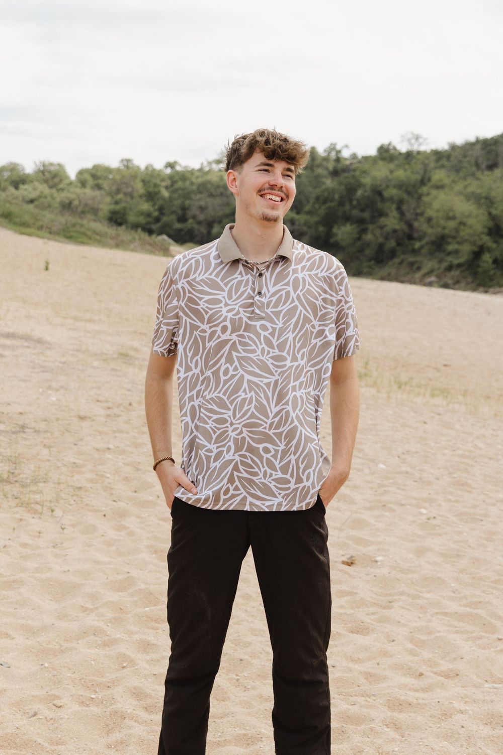 Smiling young person wearing a patterned short-sleeve shirt and dark pants stands on sandy beach with trees in background.