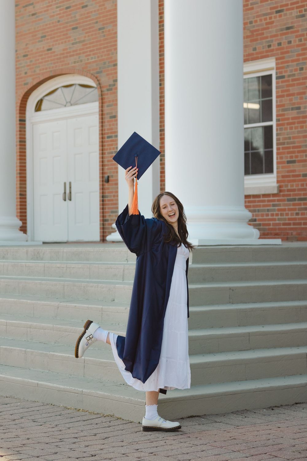 Graduate in navy cap and gown celebrates with joyful pose on brick building steps with white columns.