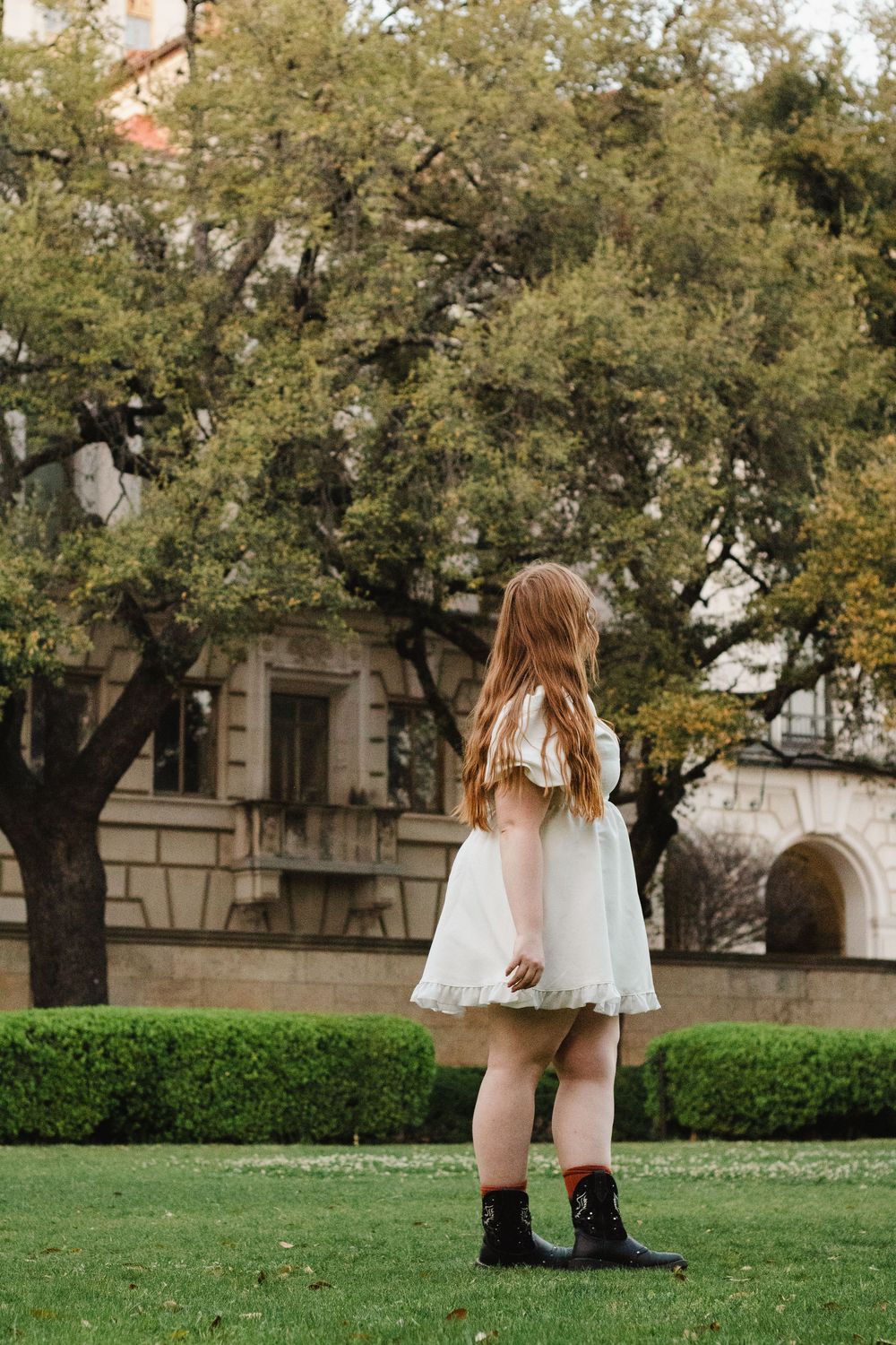 Someone in a white dress and combat boots stands on a lawn in front of historic stone buildings and trees.