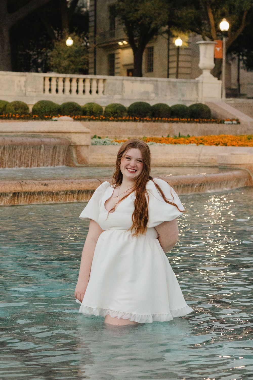 Person wearing white dress standing in fountain water at outdoor venue with landscaping and warm lighting at dusk.