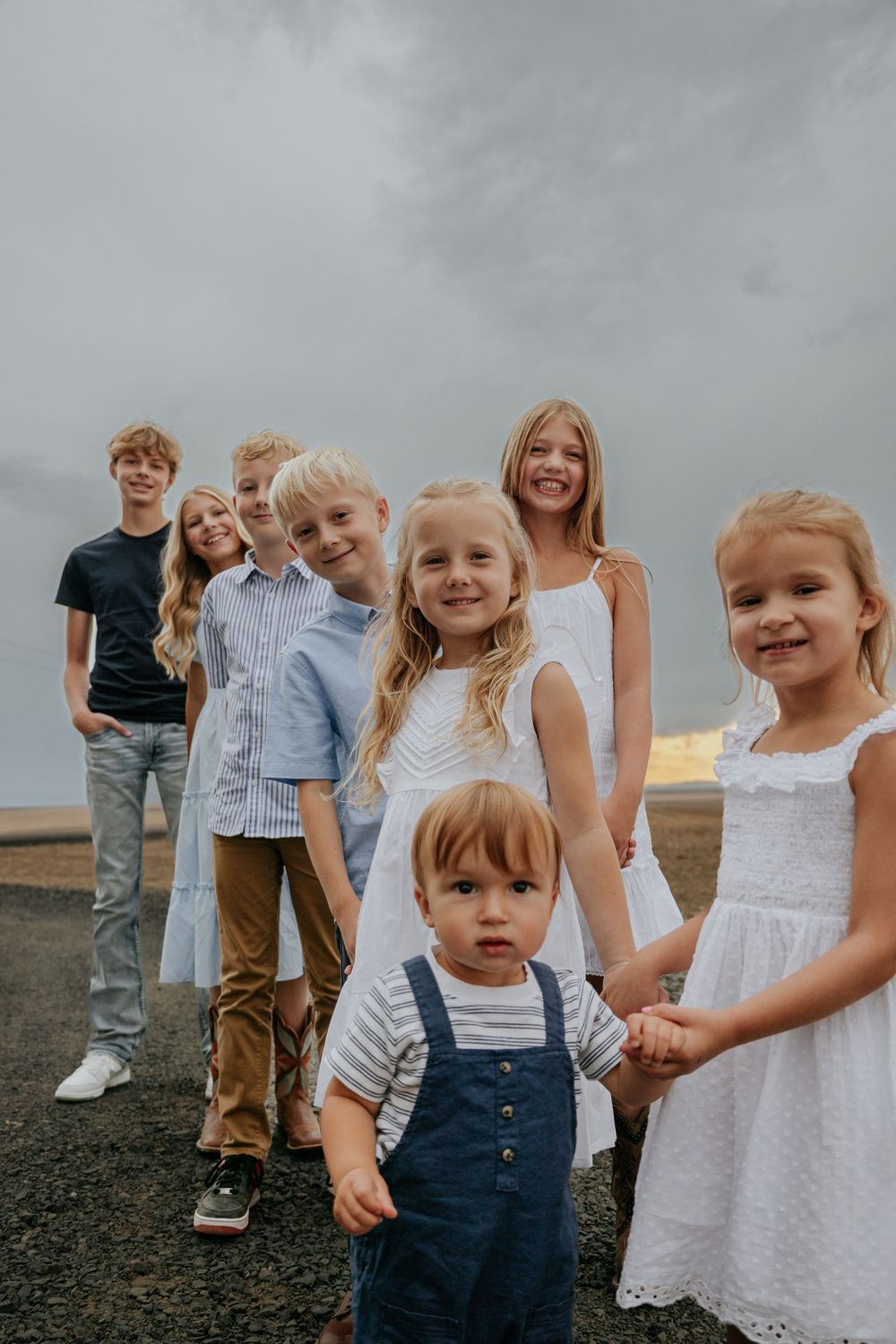 Young children in white dresses and overalls pose together against a cloudy sky.