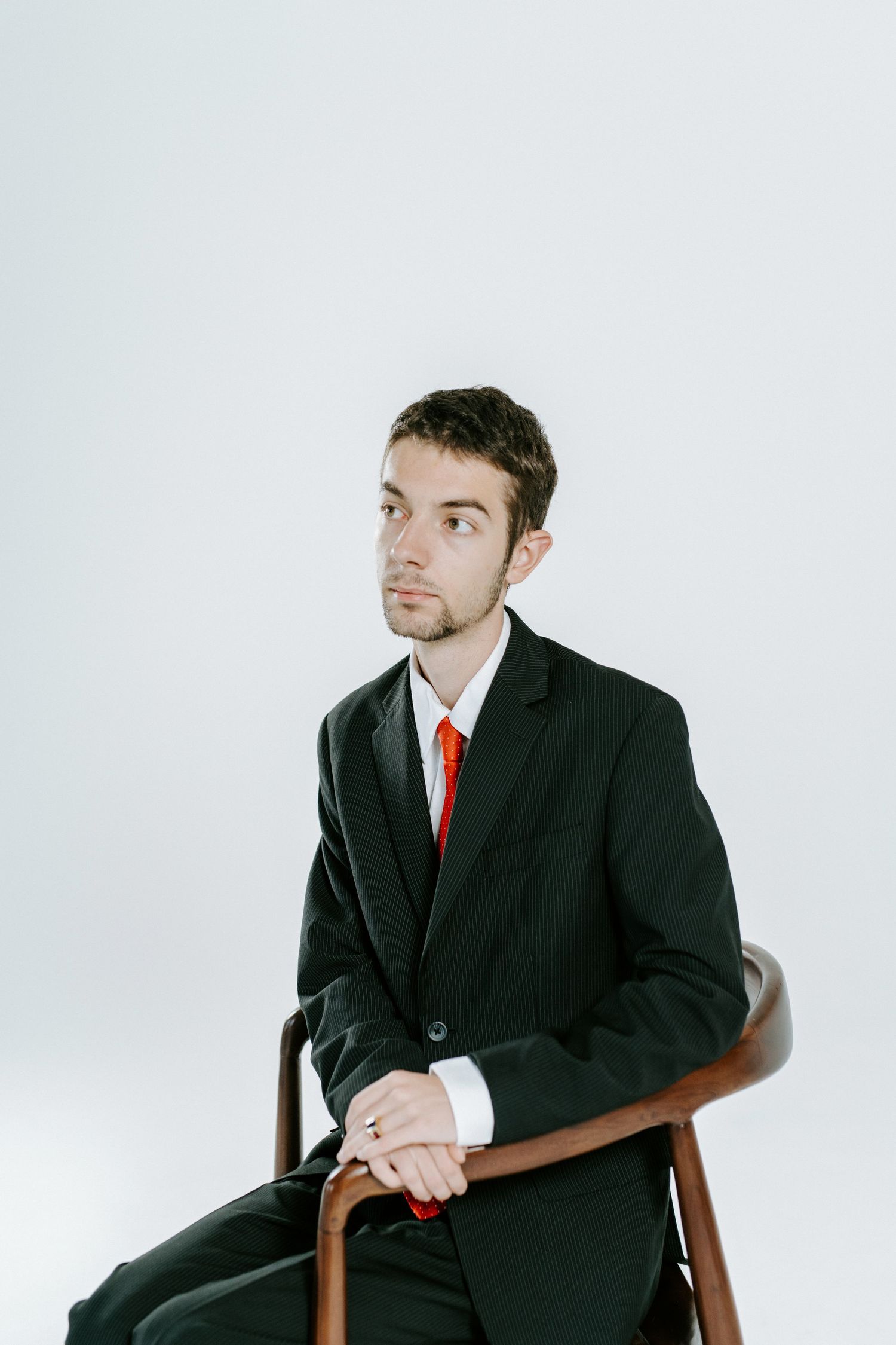 A person in a black suit and red tie sits in a wooden chair against a white background, shown from three angles.