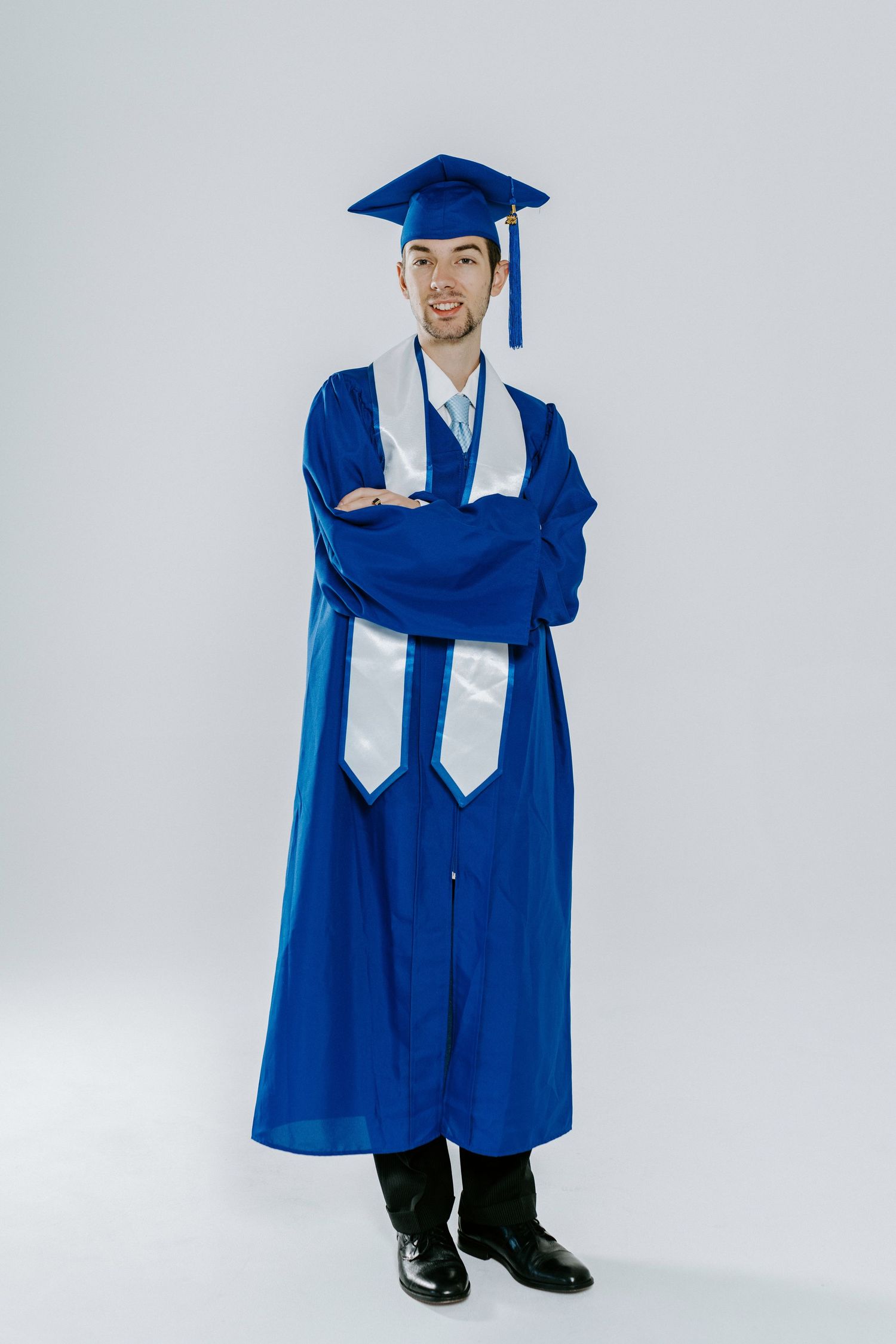 Graduate wearing traditional blue cap and gown with white stole stands confidently against light background.