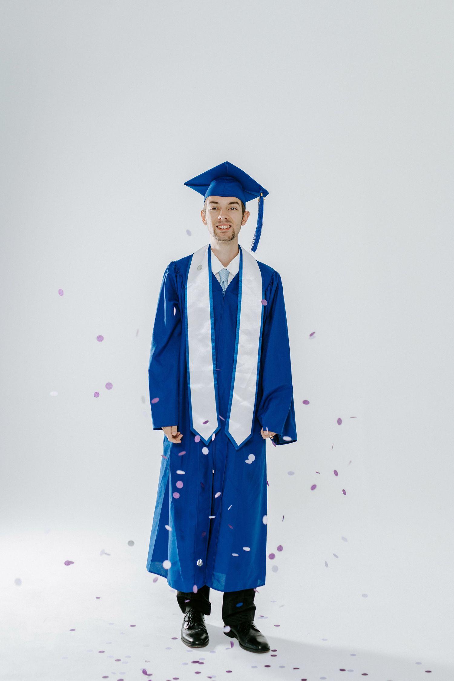 Graduate in blue cap and gown poses with confetti against white background for graduation portraits.