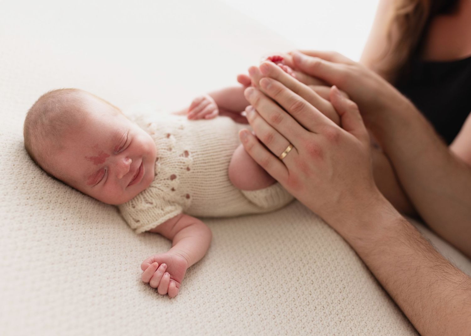 A newborn baby in a cream knit sweater lies peacefully on a white surface while gentle hands rest nearby.