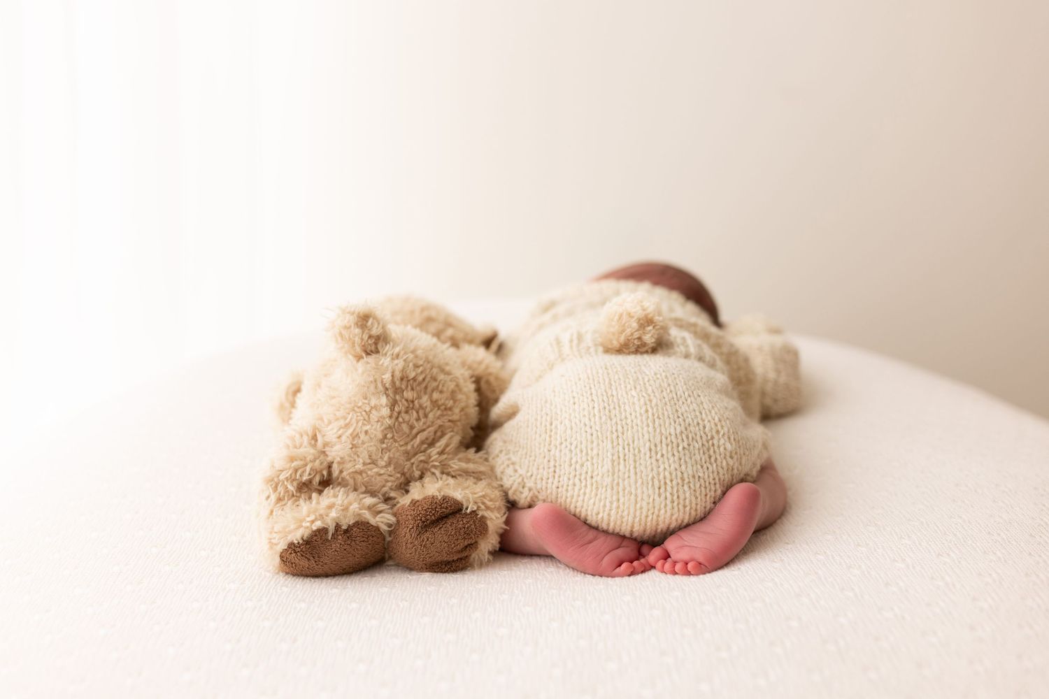 A beige teddy bear plush toy lies on a white surface in a minimalist setting.