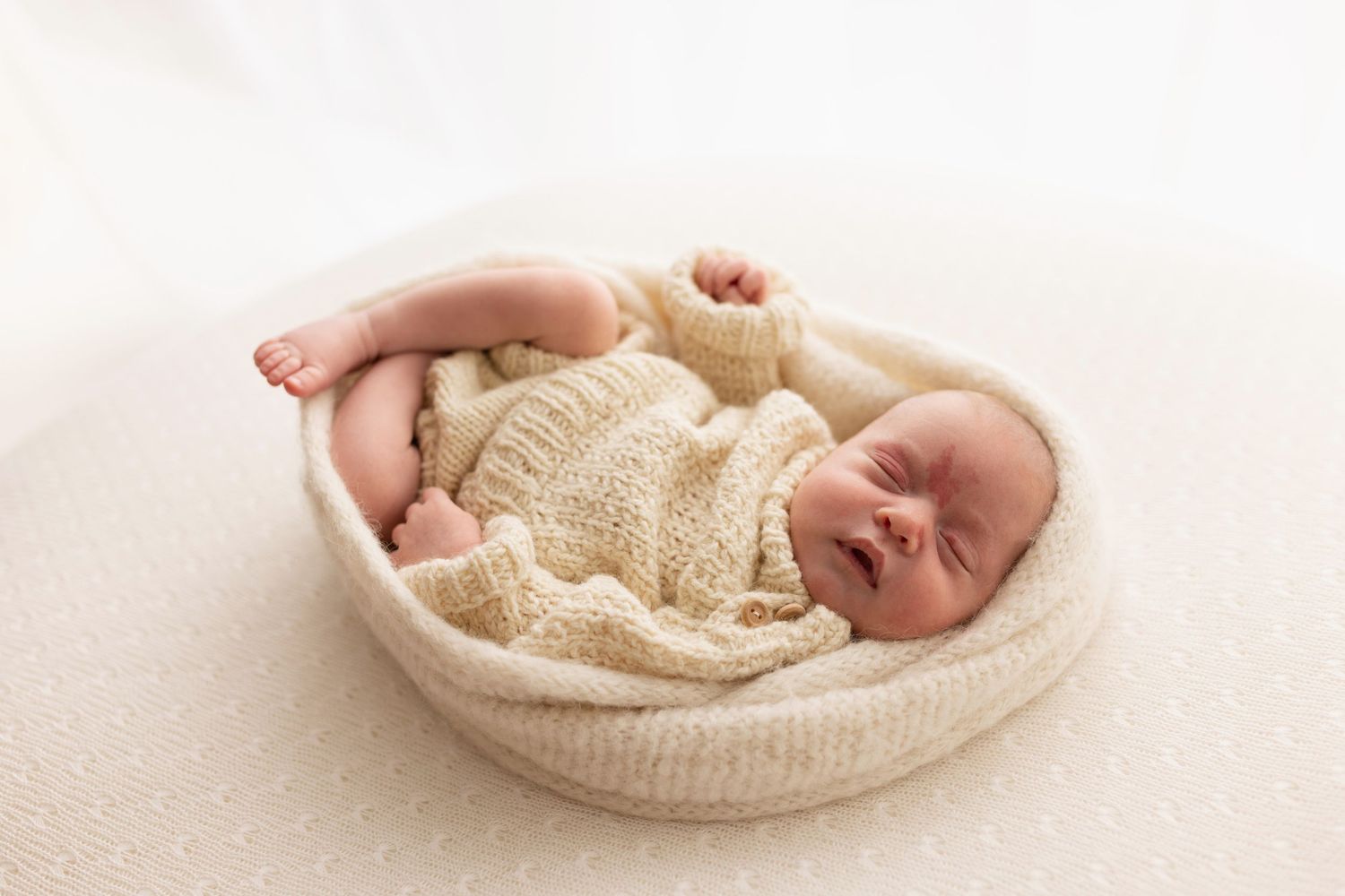 A sleeping newborn baby wrapped in cream-colored knit fabric lies curled up in a cozy round basket.