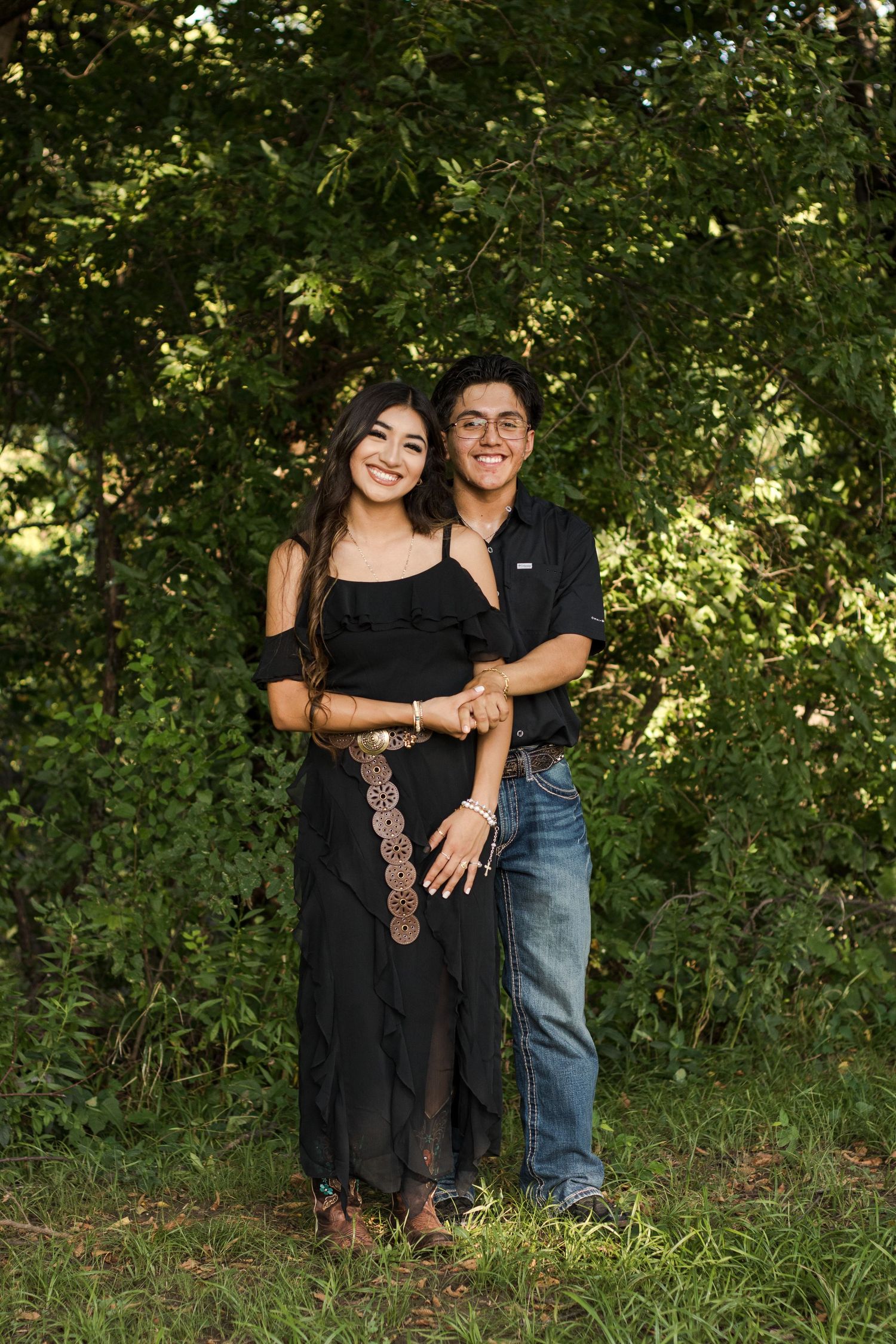 A couple wearing black and denim poses together against a lush green outdoor backdrop.