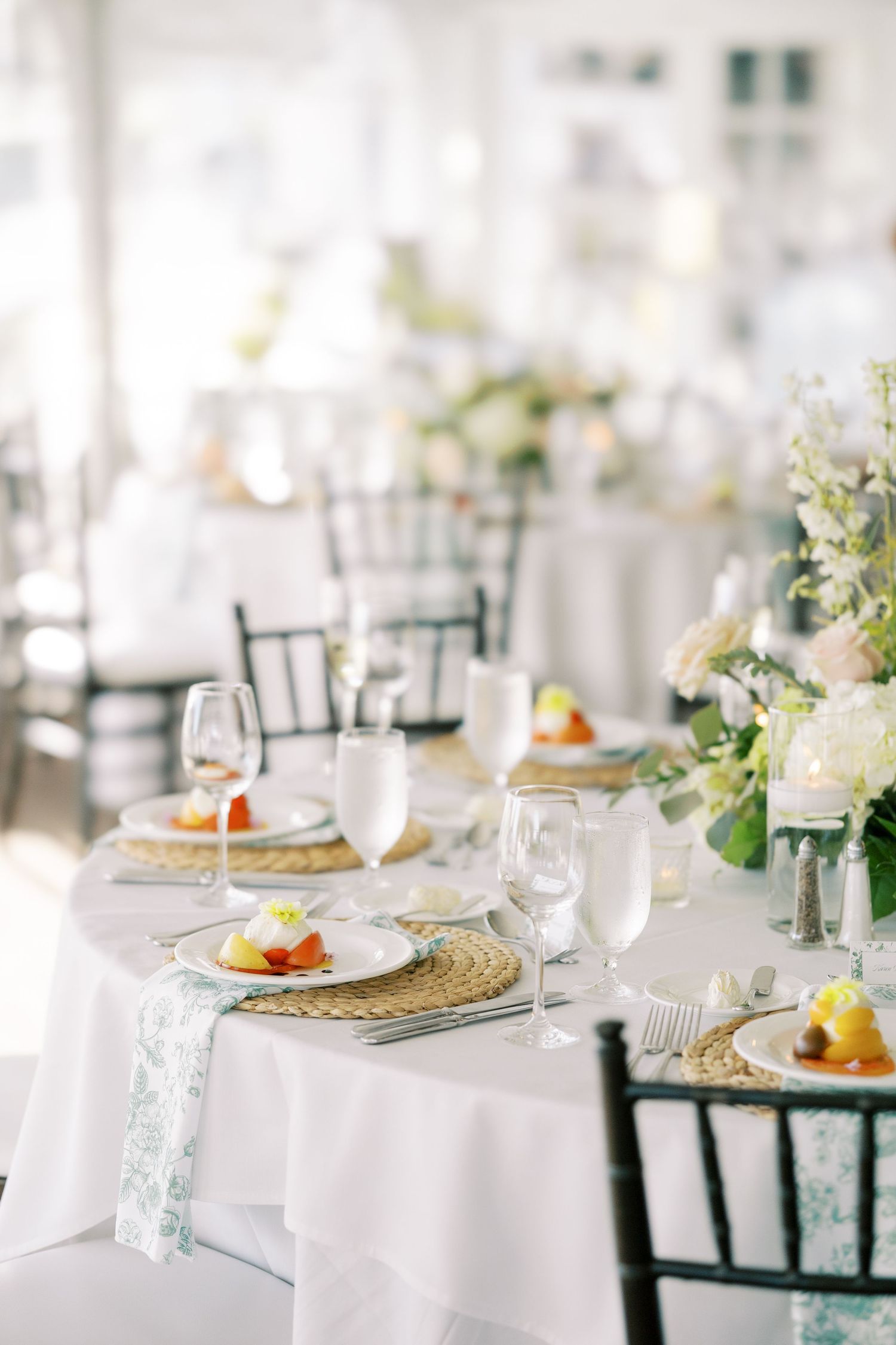 Elegant wedding reception table setting with white linens, floral centerpieces, and woven charger plates in natural light.