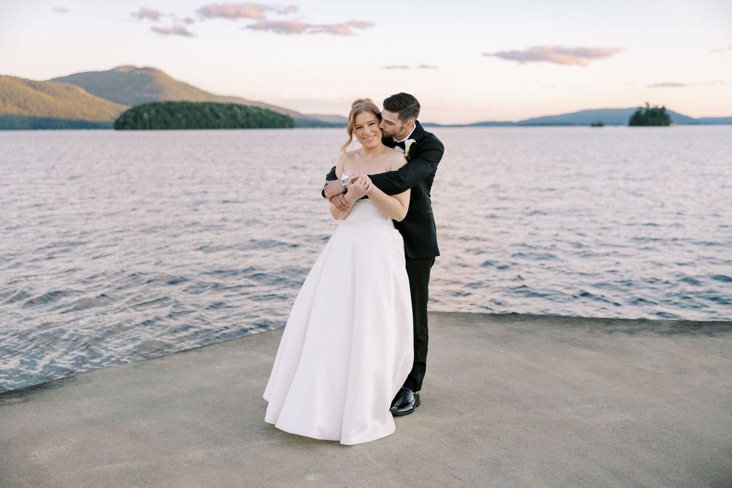 A couple in wedding attire embraces on a pier overlooking a scenic mountain lake at sunset.