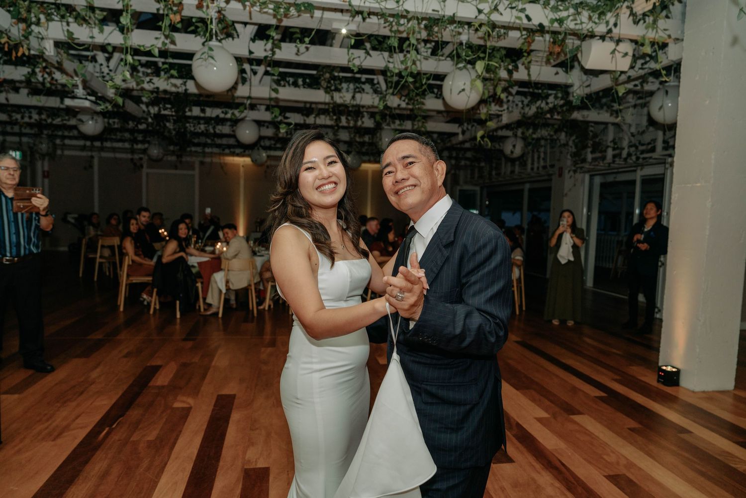 A couple dances together during their first dance at a wedding reception in a modern venue with string lights overhead.