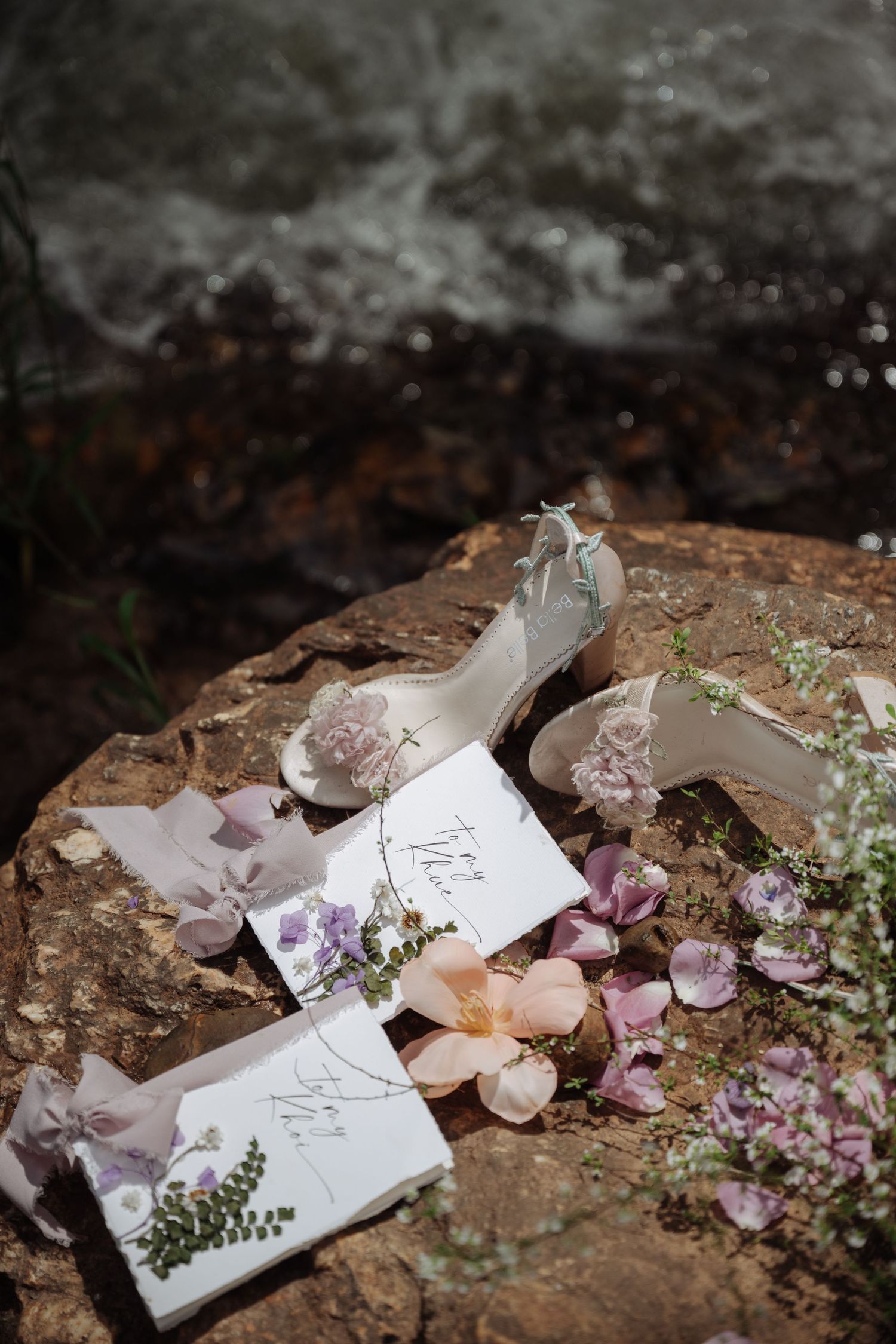 Bridal heels and floral wedding stationery arranged with scattered petals on rocky surface near water.
