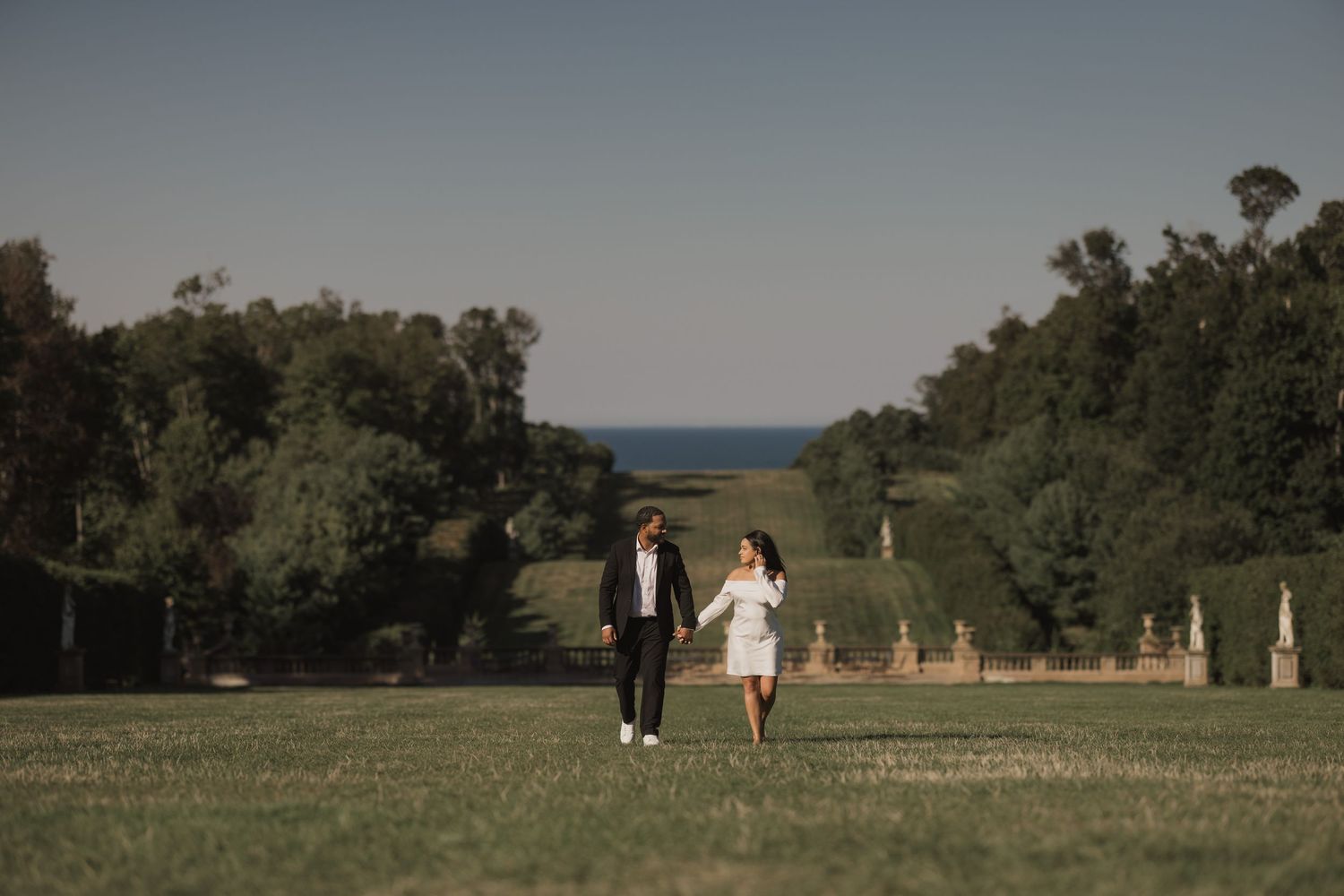 A couple walks hand in hand across a hilltop meadow with ocean views and cypress trees in the background.