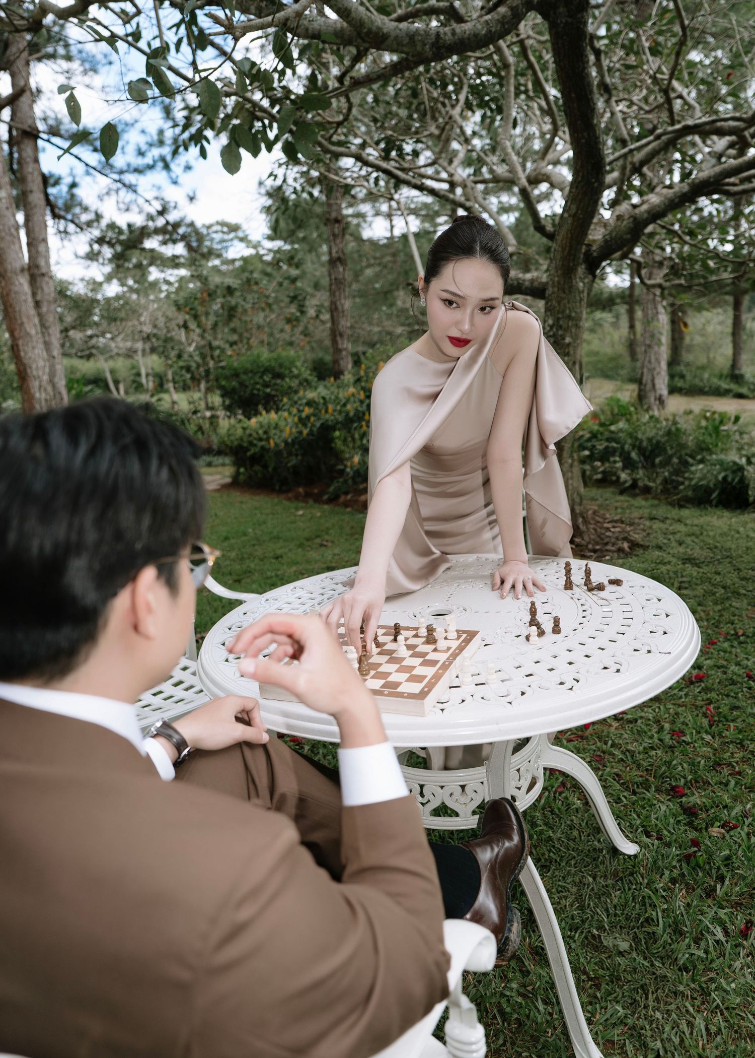 Woman in elegant beige dress leaning over chess board facing man in brown suit outdoors.