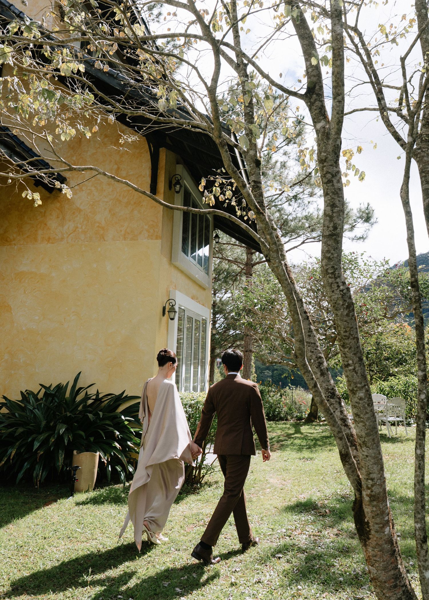 Elegant couple walking hand in hand beside a charming yellow building surrounded by trees.