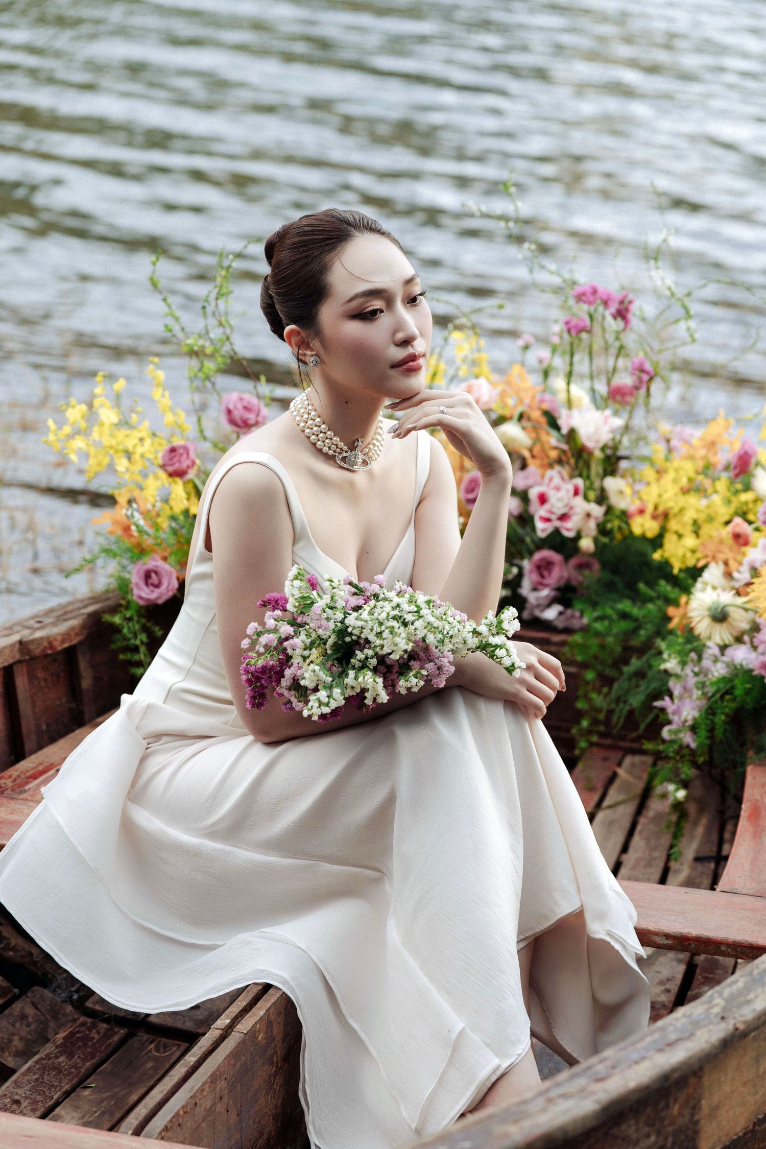 Elegant bride in white dress holding wildflowers, seated in a wooden boat surrounded by colorful blooms by a lake.
