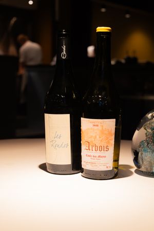 Wine bottles including Arbois label displayed on white surface in dim lighting.