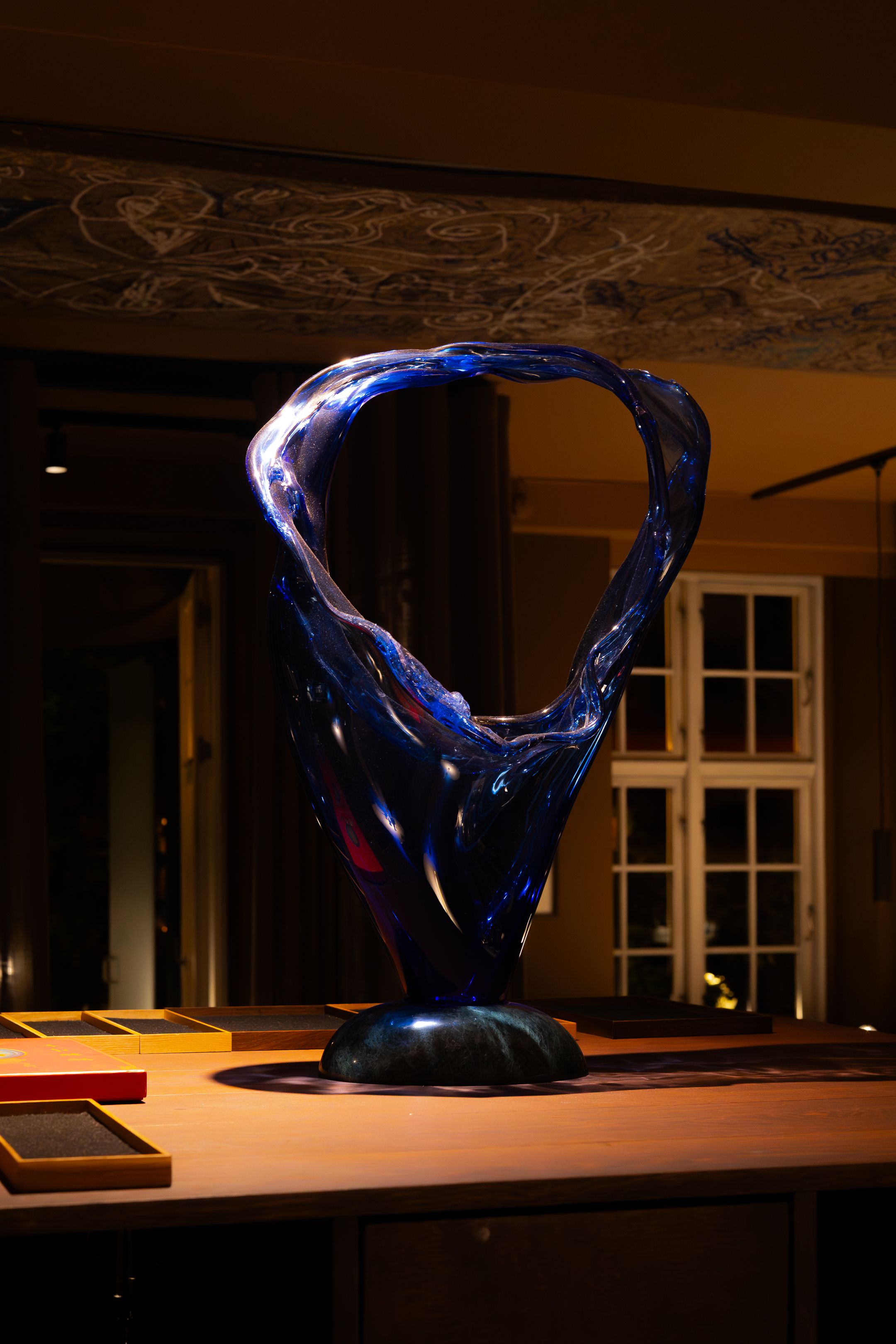 A dark blue glass sculpture sits illuminated on a wooden shelf in dramatic low lighting creating an artistic display.