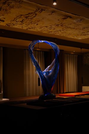 A modern blue glass sculpture illuminated in dim lighting sits atop a dark surface with decorative ceiling details visible above.