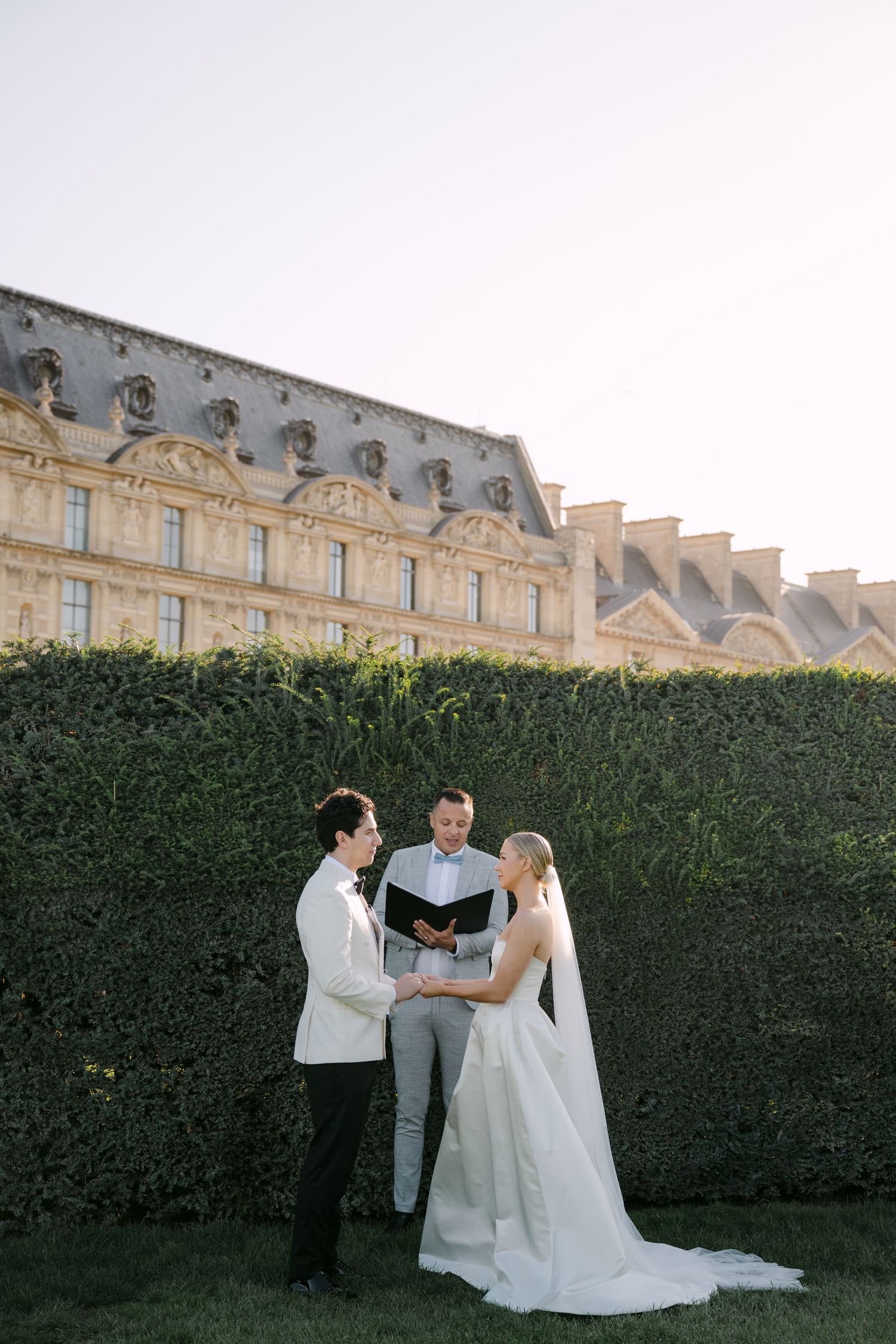 Wedding party poses together in front of elegant French architecture and manicured hedges.