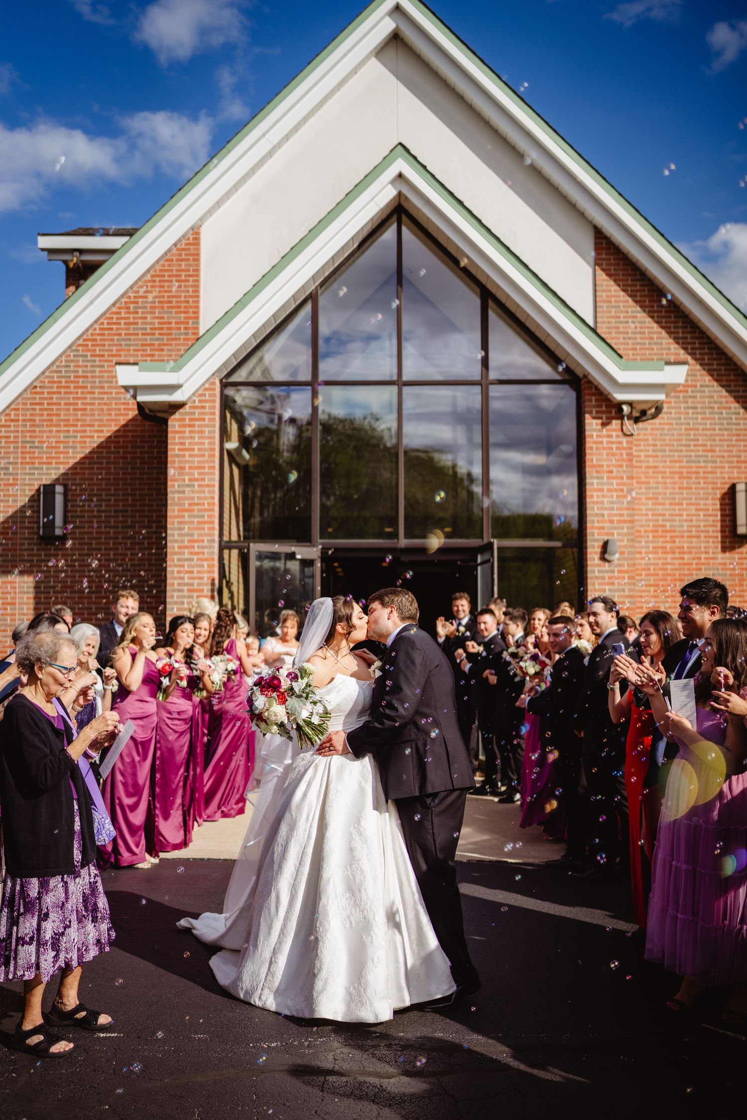 Outdoor wedding celebration with people gathered in front of a brick church on a sunny day with blue skies.