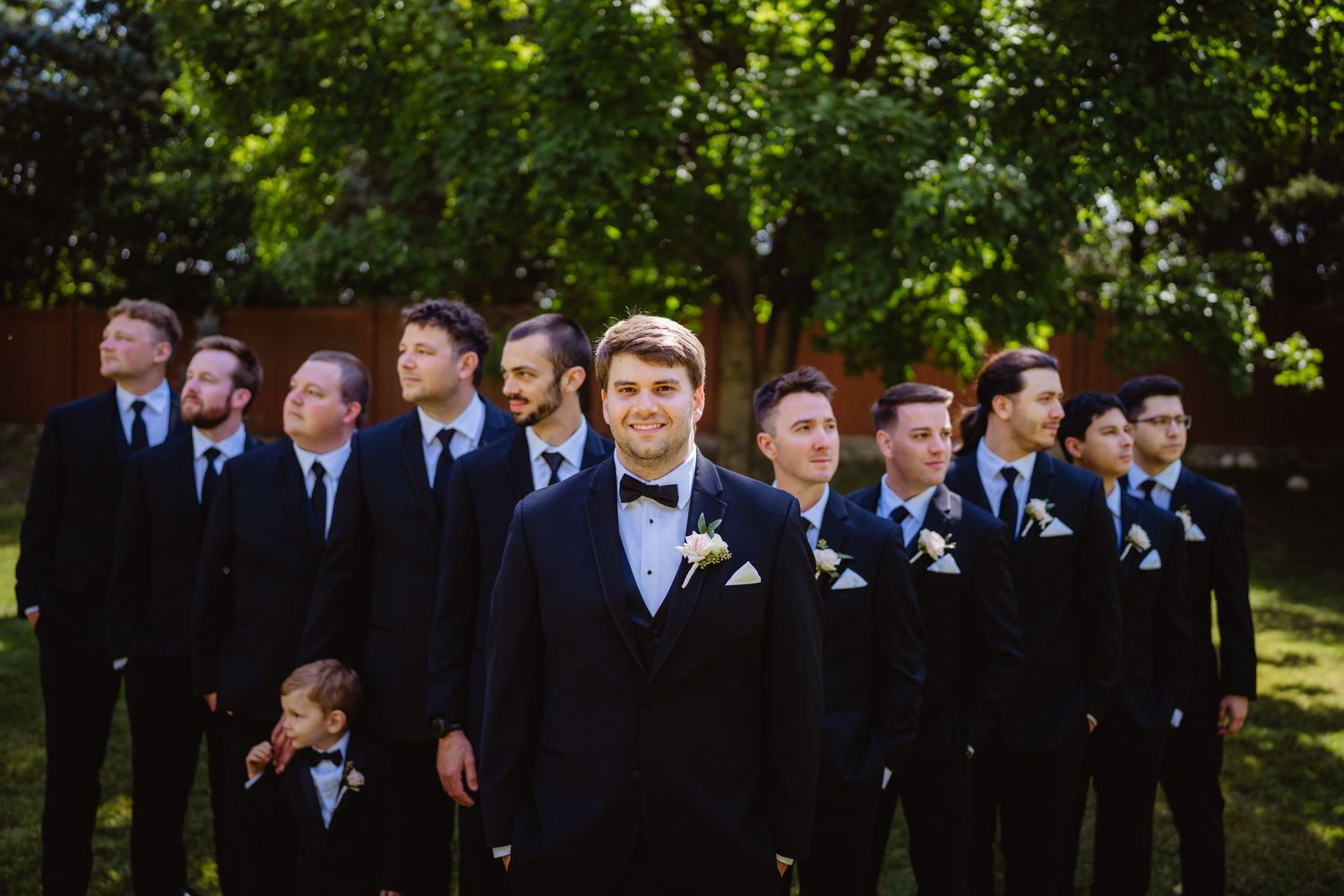 A wedding party in navy suits and light blue ties poses outdoors among trees and sunlight.