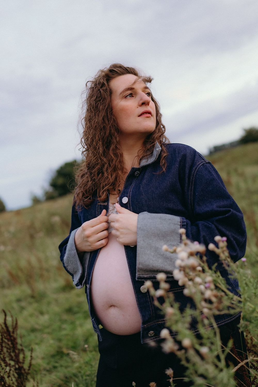 Person in denim jacket posing in a field of wildflowers during a maternity photoshoot.