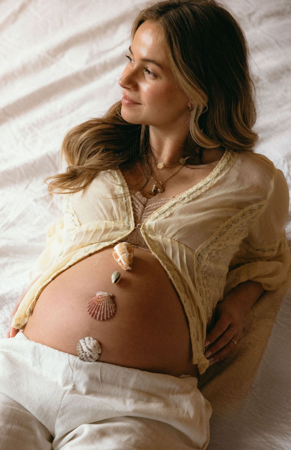 Maternity portrait with person wearing cream lace top and seashell necklace on white bedding.