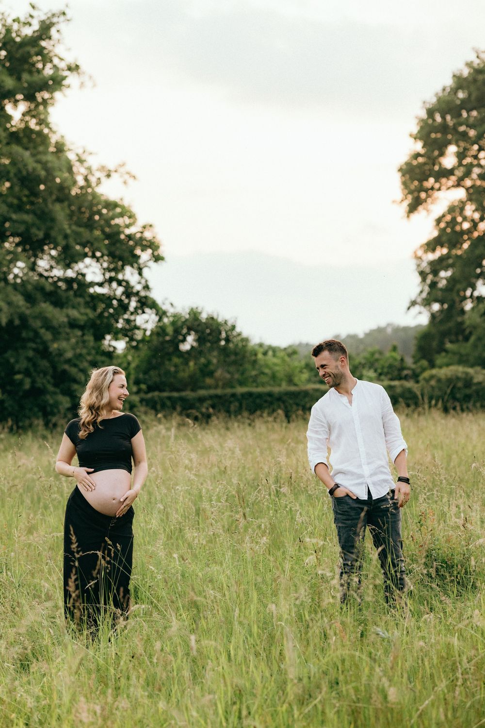 Couple poses playfully in a scenic meadow with trees in the background.
