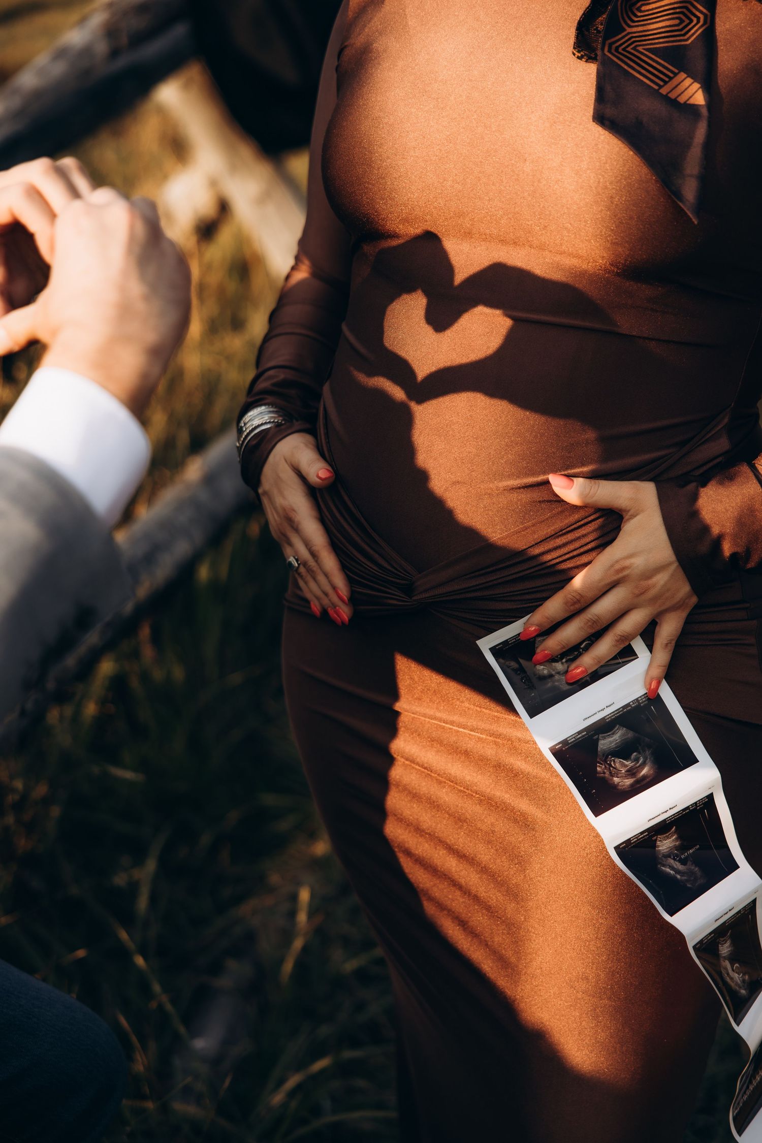 Hands form a heart shape shadow on pregnant belly while holding ultrasound photos in warm sunlight.