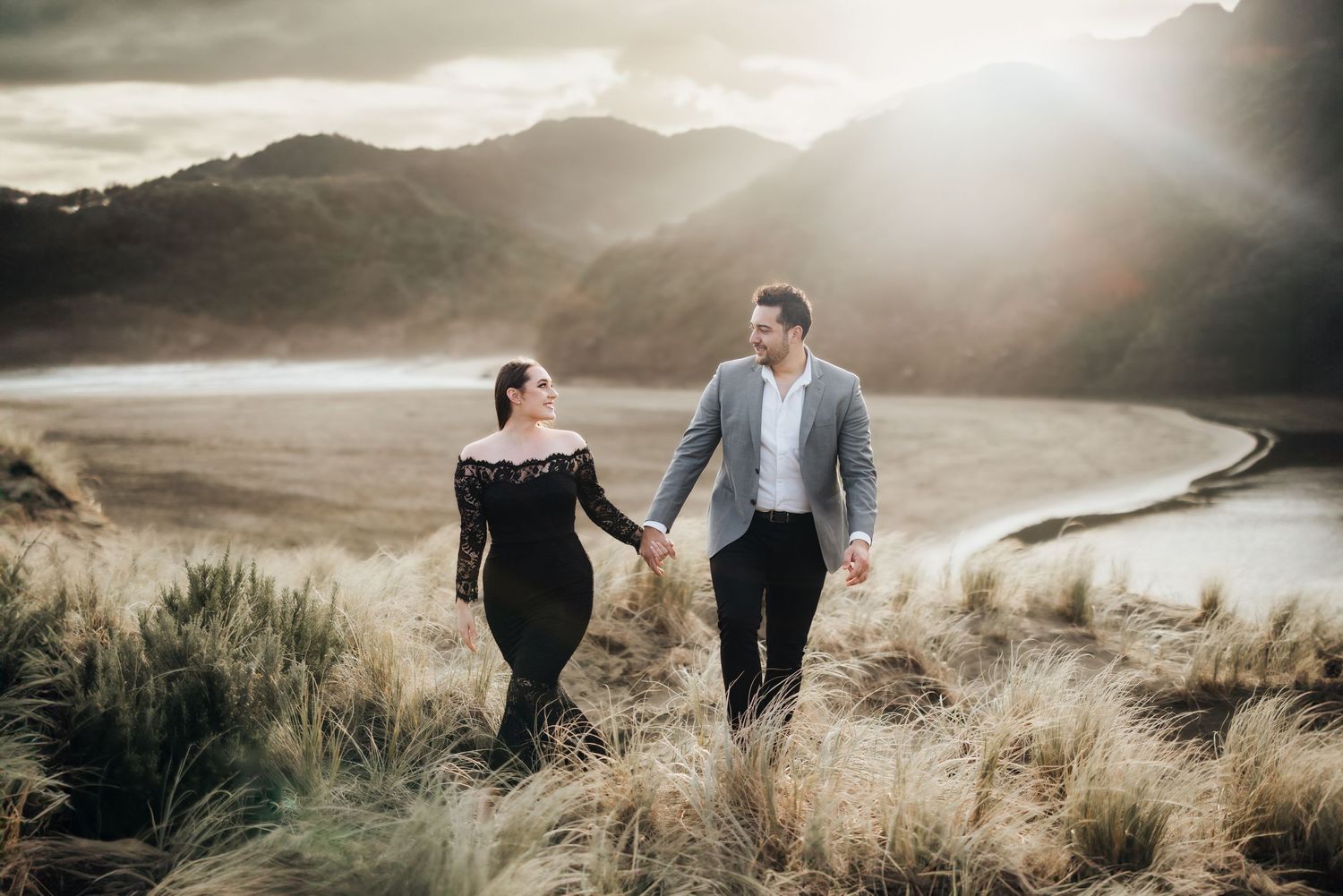 A couple walking hand in hand along a hilltop path at sunset overlooking a scenic mountain reservoir.