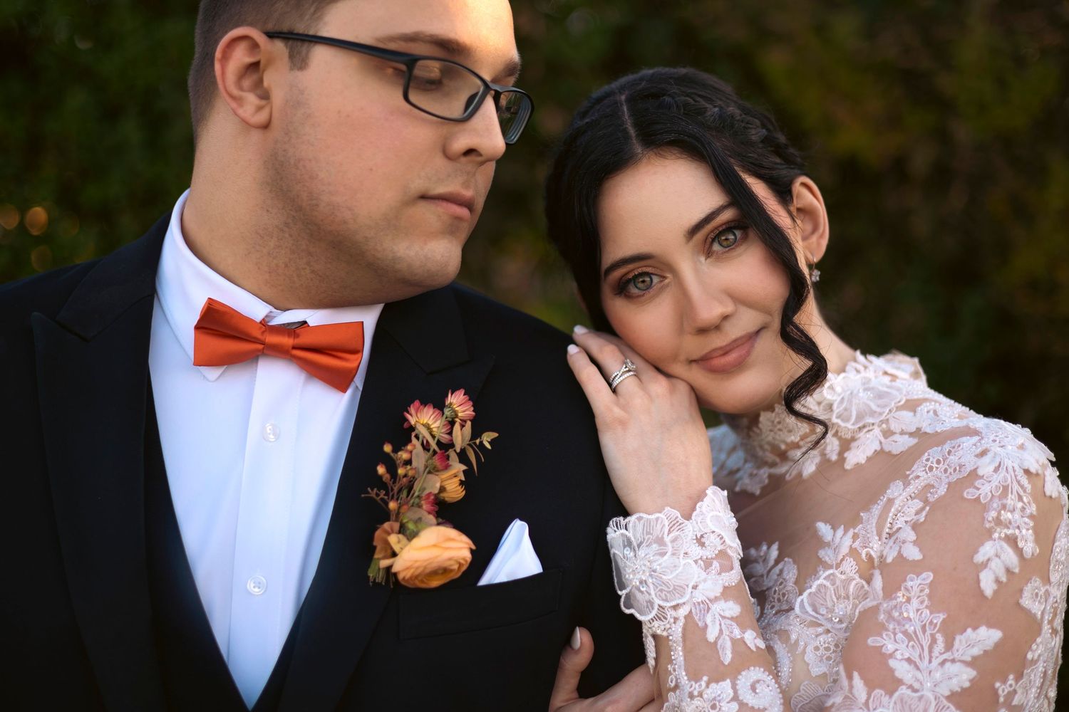 Elegant bride in lace long sleeve dress poses with groom wearing black tuxedo and red bow tie during outdoor wedding.